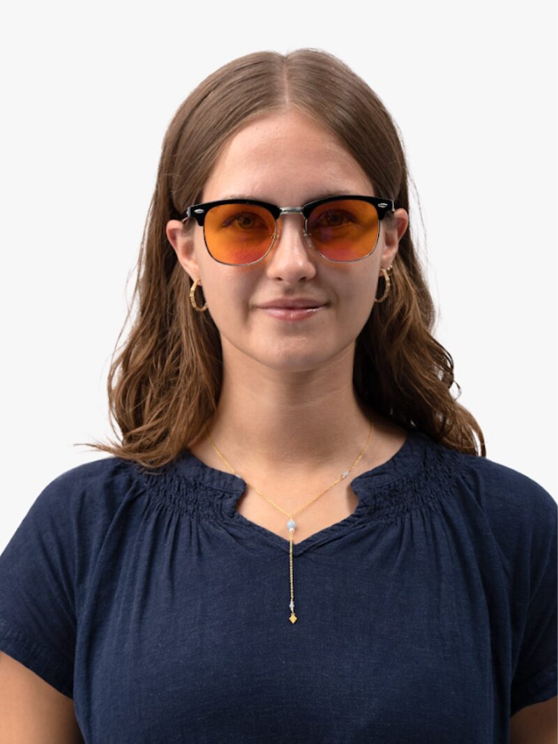Junge Frau mit orange getönter Brille, goldenen Creolen und feiner Goldkette mit Anhänger, dunkelblaue Bluse