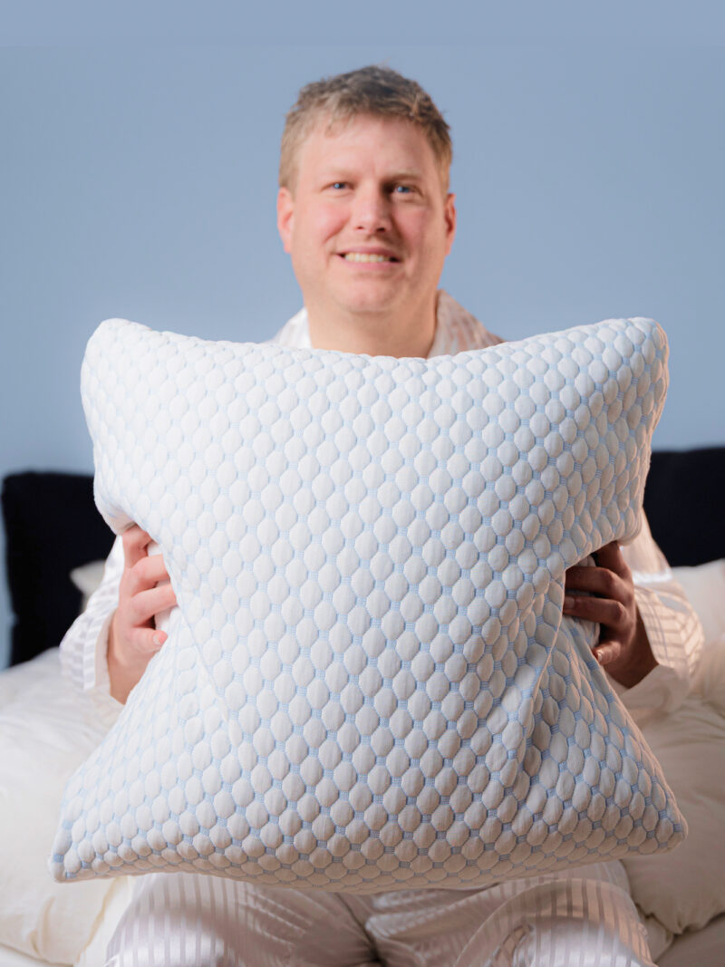Mann im Pyjama hält ein großes weißes Kissen mit gestepptem Sechseckmuster auf dem Bett