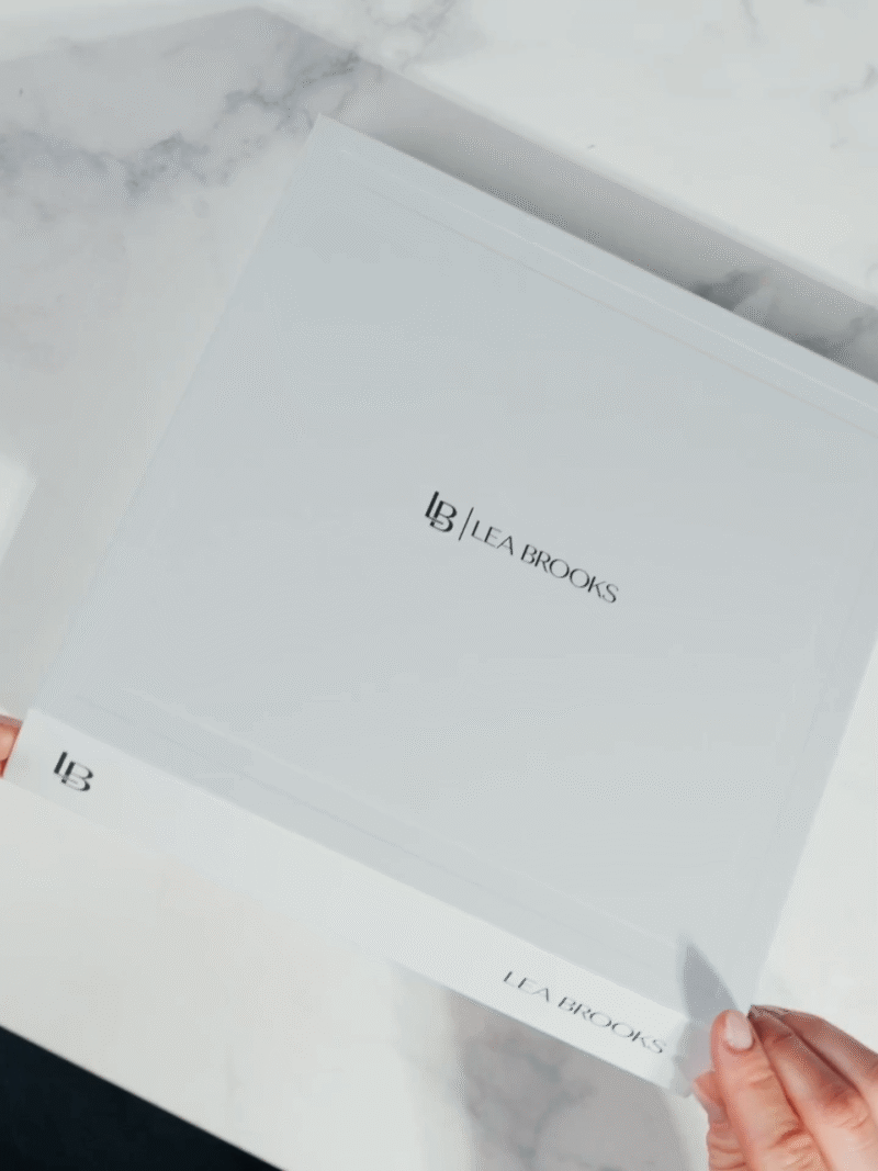 Weiße Lea Brooks Geschenkbox mit schwarzem Logo, Hände öffnen den Deckel auf einem Marmortisch
